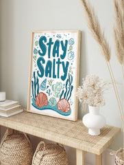 Stay Salty typography art print with teal text, orange and blue seashells, green seaweed on, in an alternate framed mockup