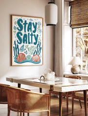 Stay Salty typography art print with teal text, orange and blue seashells, green seaweed on white, in a styled room mockup