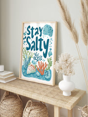 Stay Salty typography print with blue text, orange coral, green seaweed, and seashells on a, in an alternate framed mockup