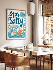 Stay Salty typography print with blue text, orange coral, green seaweed, and seashells on a white, in a styled room mockup
