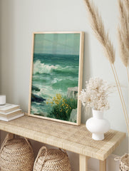 Painting of stormy green sea with a large white wave crashing on rocks and yellow flowers, in an alternate framed mockup