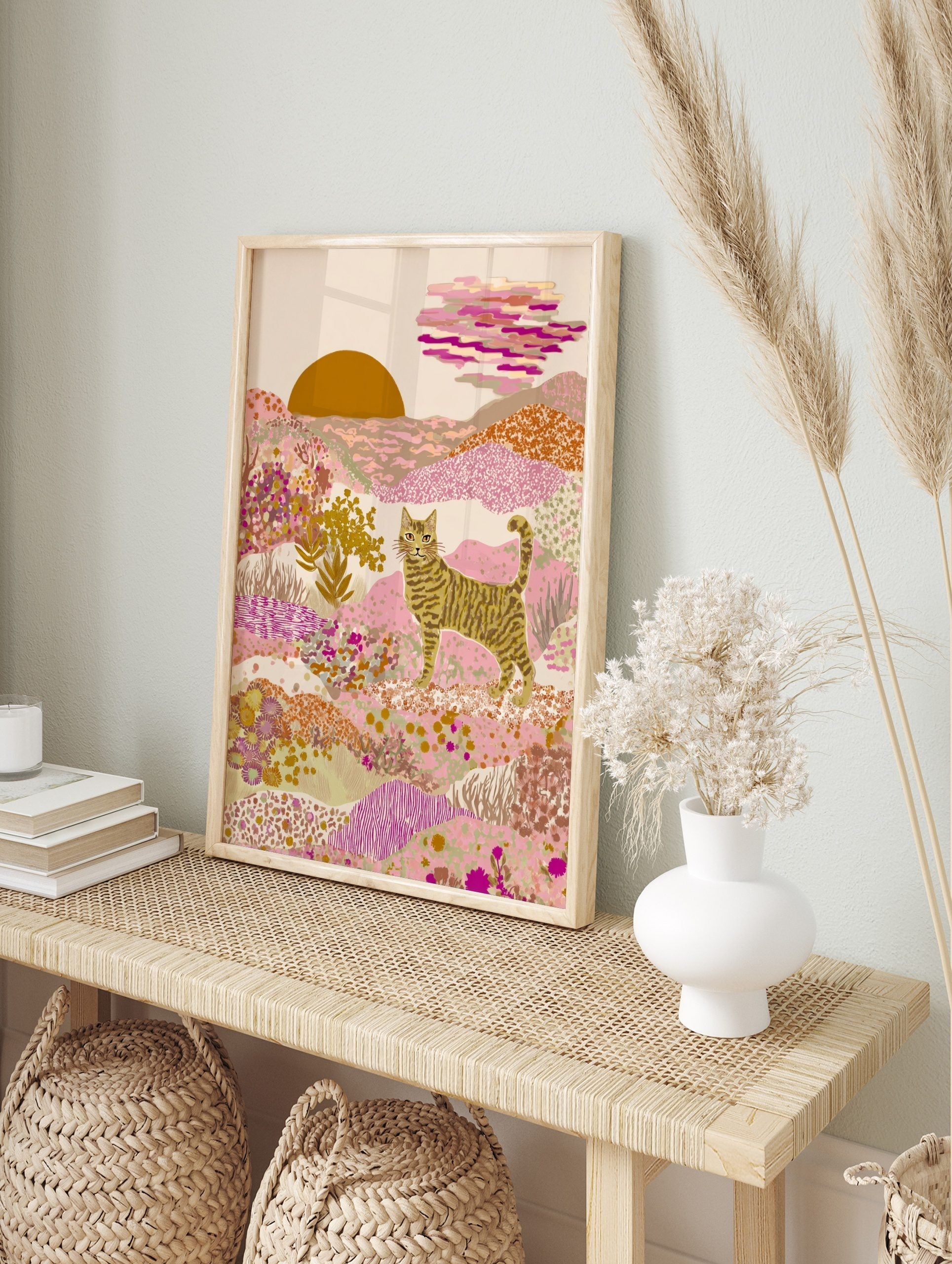 Illustrative striped cat in a pink and gold desert landscape with patterned hills and a, in an alternate framed mockup