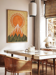Illustrative orange sun with jagged rays rising behind a snow-capped mountain wall art print, in a styled room mockup