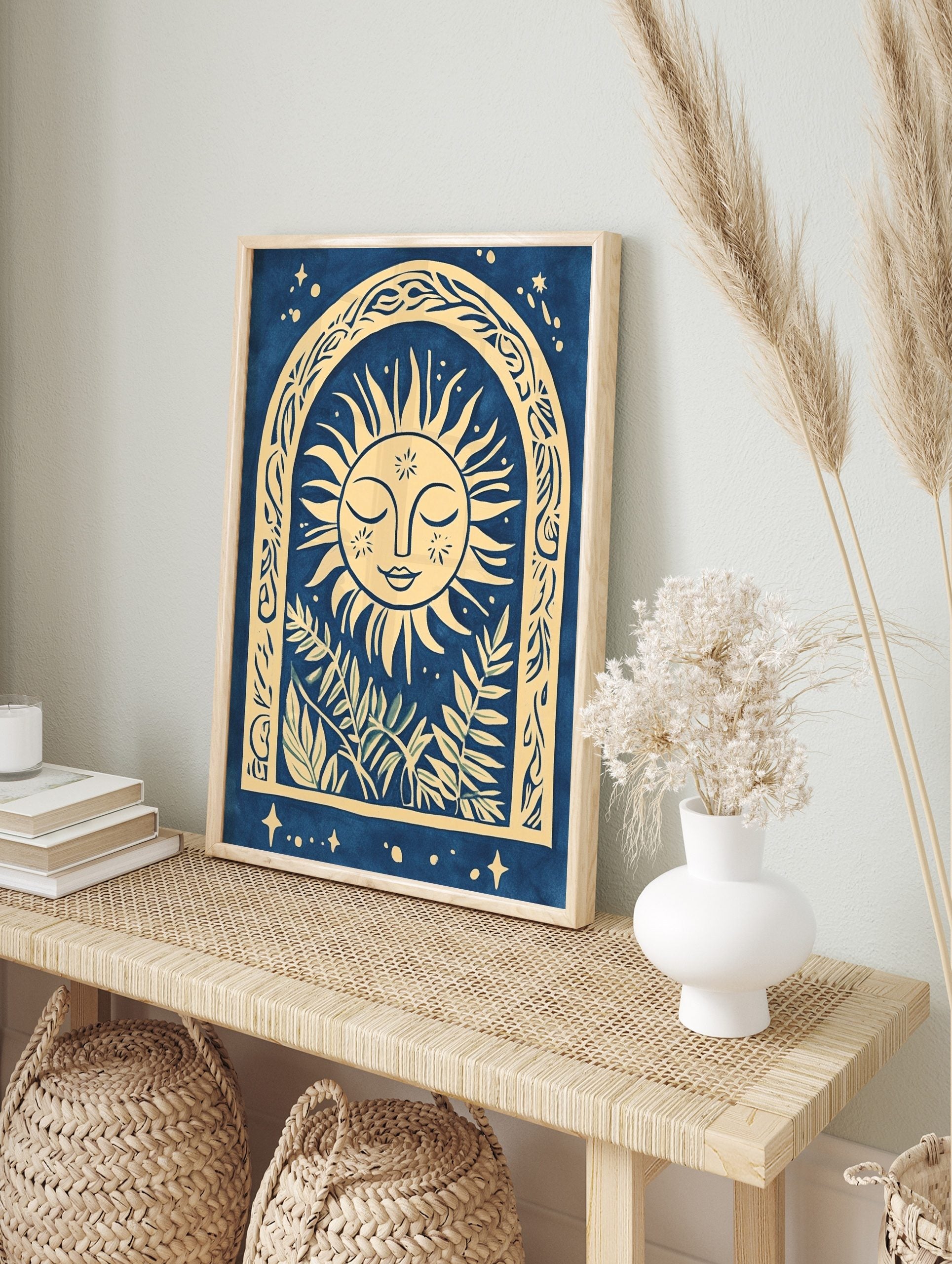 Illustrative sun face with closed eyes in an ornate arch with botanical leaves, blue and, in an alternate framed mockup