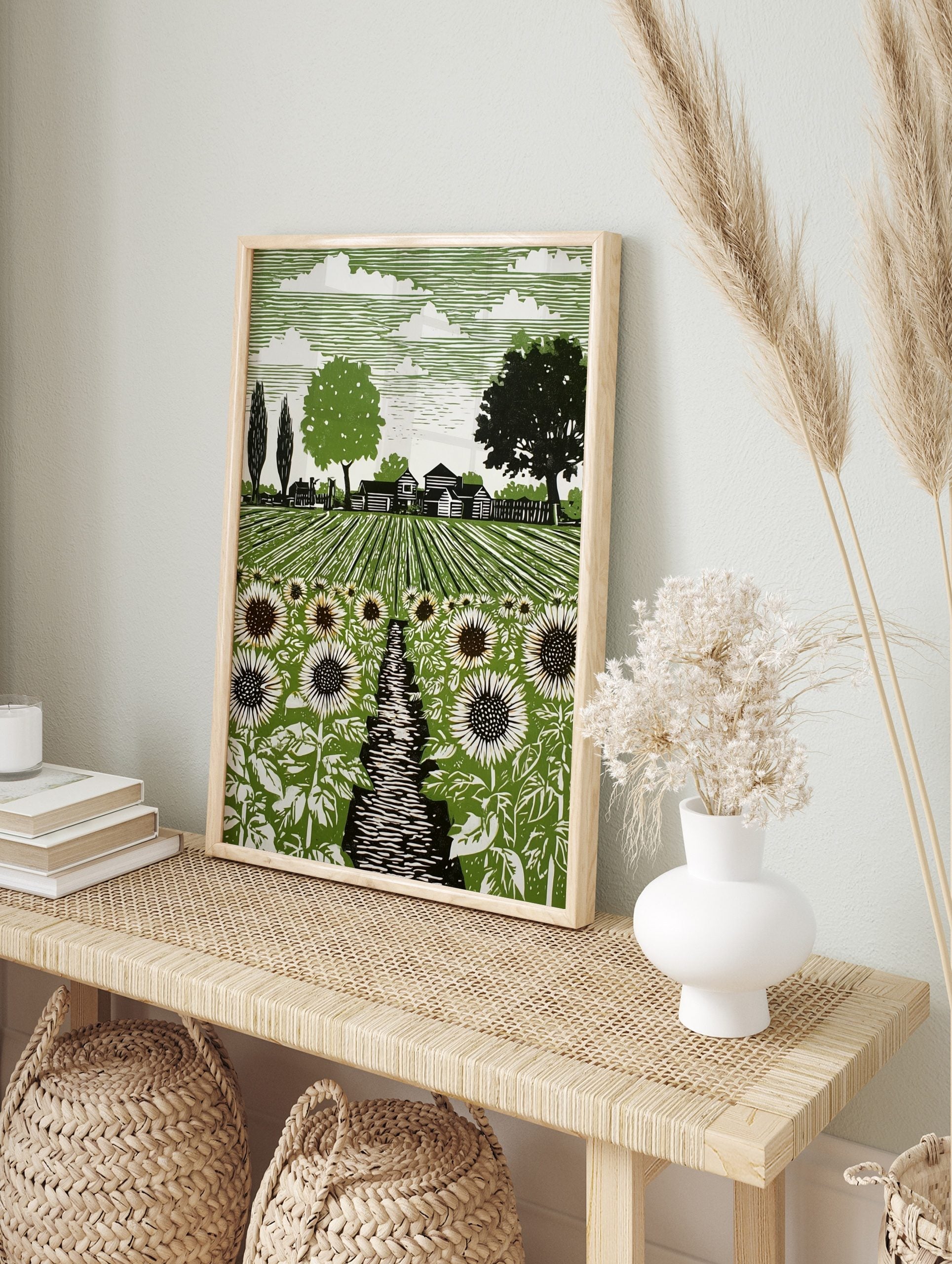 Linocut style art print of a sunflower field with a path, farmhouses, and trees under a, in an alternate framed mockup