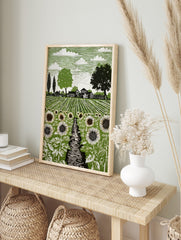 Linocut style art print of a sunflower field with a path, farmhouses, and trees under a, in an alternate framed mockup