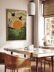 Linocut-style art print of two birds flying over an orange sun, green fields, and tall foreground, in a styled room mockup
