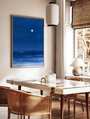 Minimalist blue night sea art print featuring a lone surfer gliding under a large full moon, in a styled room mockup