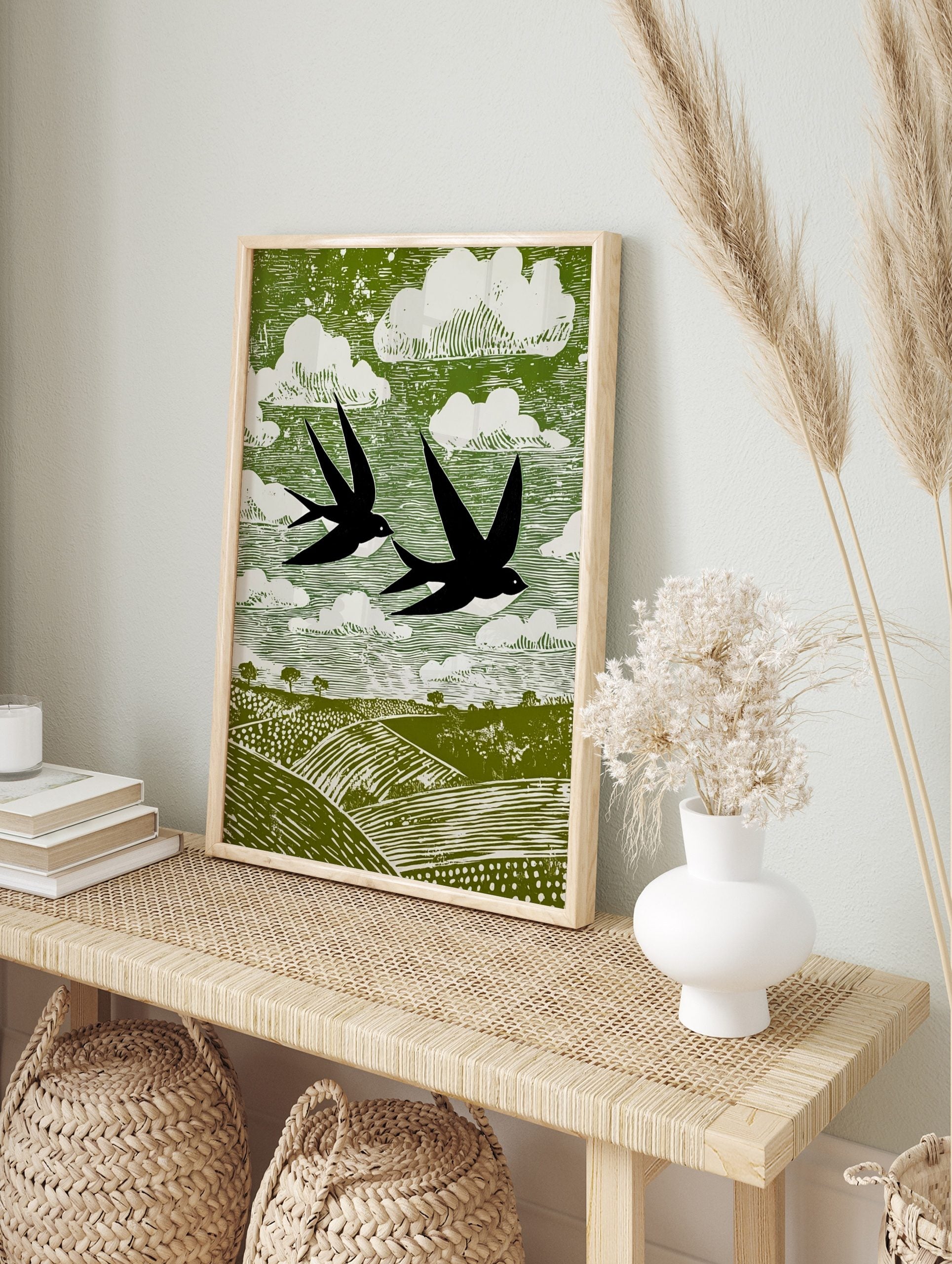 Stylised linocut print of two black swallows flying over green fields and white clouds in a, in an alternate framed mockup