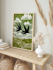 Stylised linocut print of two black swallows flying over green fields and white clouds in a, in an alternate framed mockup