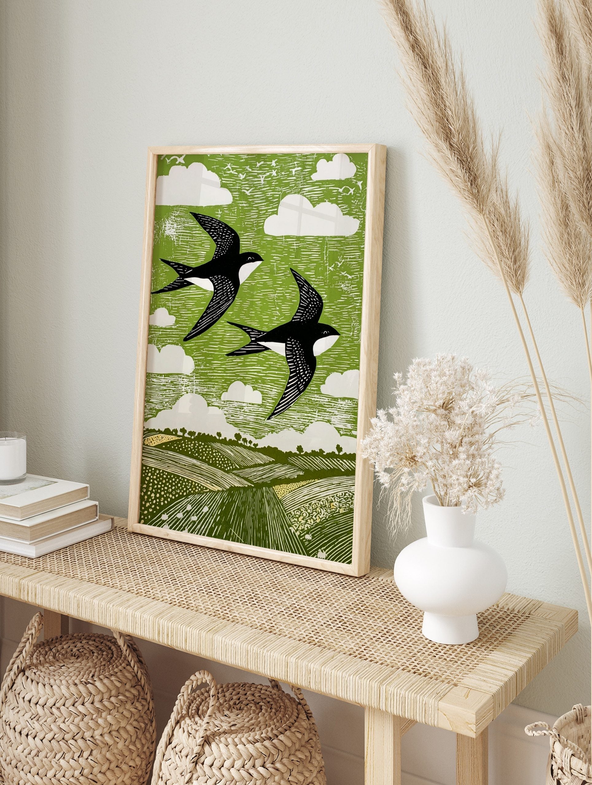 Linocut style print of two black and white swallows flying over green fields and sky with, in an alternate framed mockup
