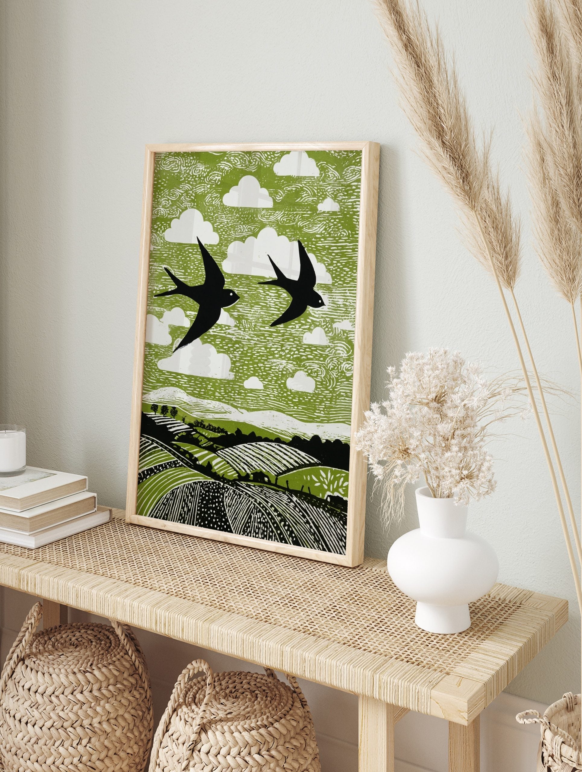 Linocut style print of two black swallows flying over green fields with white clouds, in an alternate framed mockup