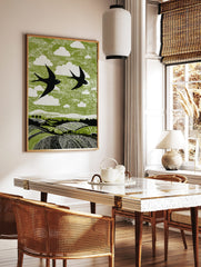 Linocut style print of two black swallows flying over green fields with white clouds, in a styled room mockup