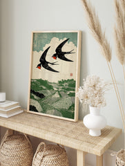 Woodcut print of two black and red swallows flying over a green rolling landscape with fields, in an alternate framed mockup