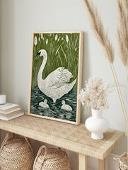 White swan and two cygnets swimming in green water with tall reeds and cattails in, in an alternate framed mockup