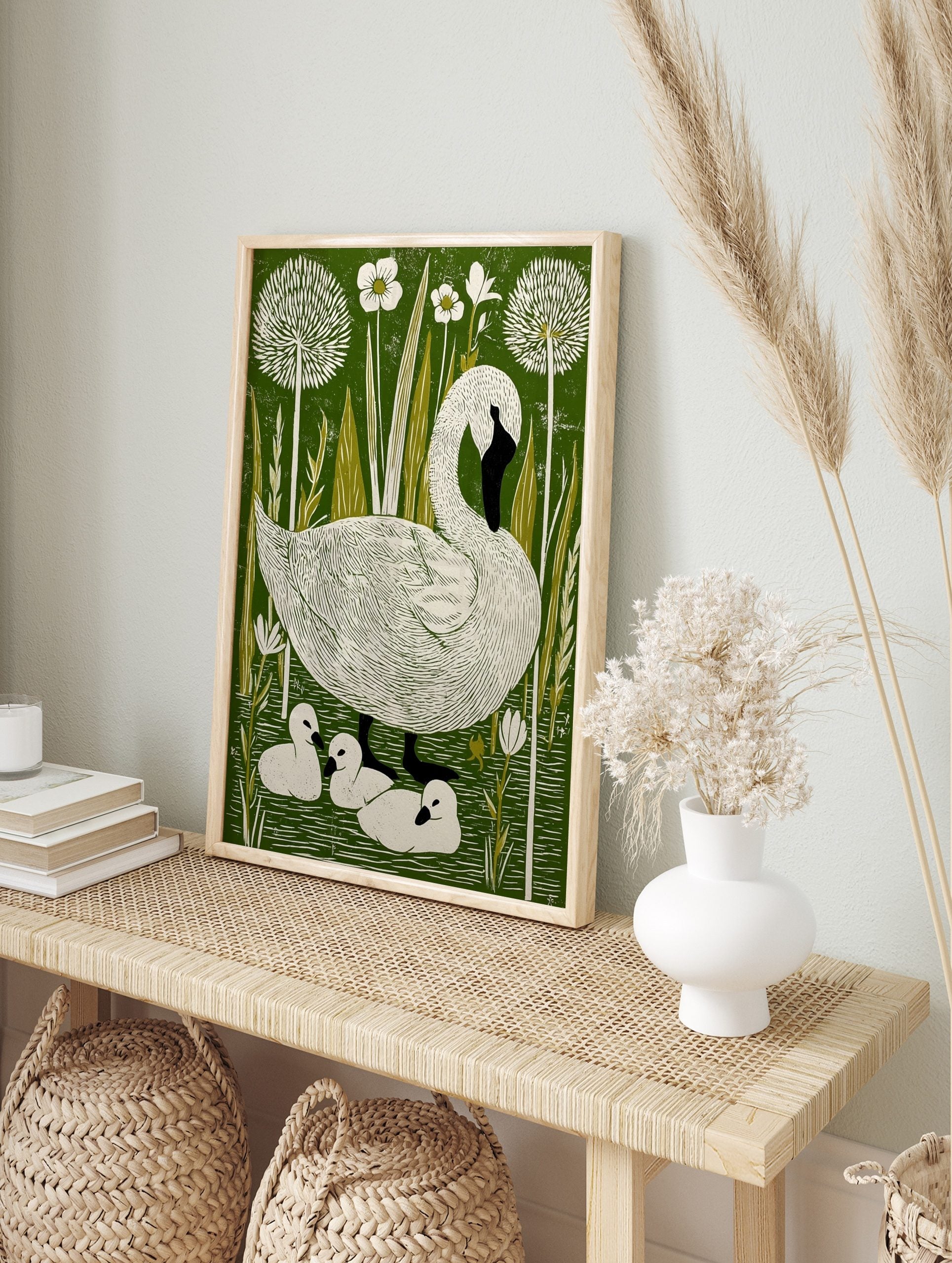 Illustrative linocut-style print of a white swan and three cygnets in a green pond with white, in an alternate framed mockup