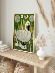 Illustrative linocut-style print of a white swan and three cygnets in a green pond with white, in an alternate framed mockup