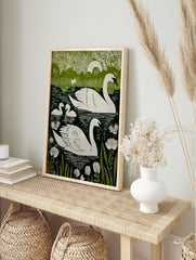 Green and white linocut-style print of two swans and two cygnets swimming in a lake with, in an alternate framed mockup