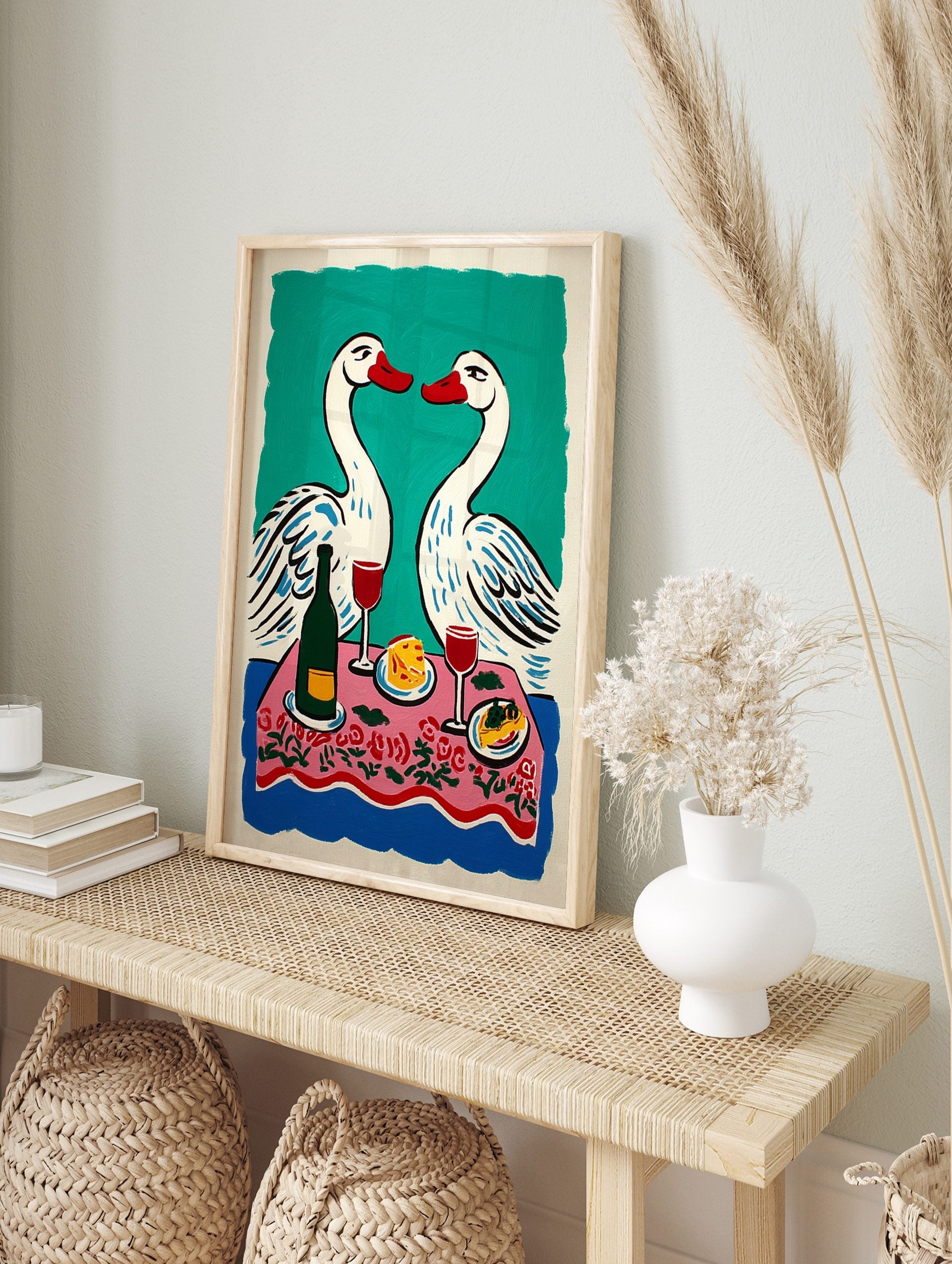 Illustrative print of two white swans with red beaks over a pink floral dinner table with, in an alternate framed mockup
