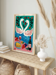 Illustrative art print of two white swans forming a heart, a blue lake, red flowers, and a, in an alternate framed mockup
