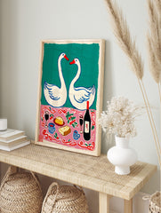 Illustrative print of two white swans forming a heart above a pink tablecloth with wine, in an alternate framed mockup
