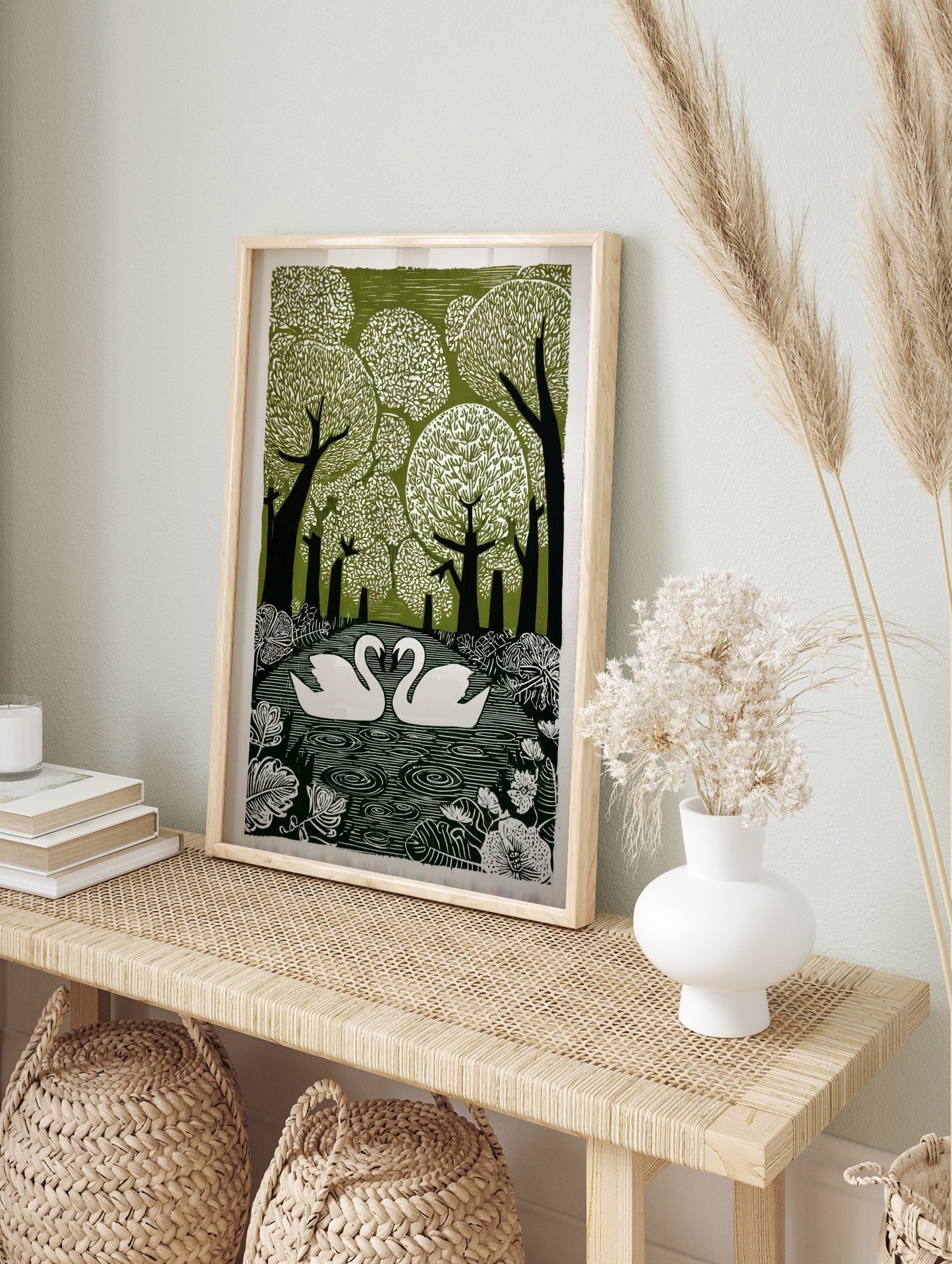 Linocut print of two white swans on water, surrounded by green trees and botanical leaves, in an alternate framed mockup