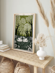 Linocut print of two white swans on water, surrounded by green trees and botanical leaves, in an alternate framed mockup