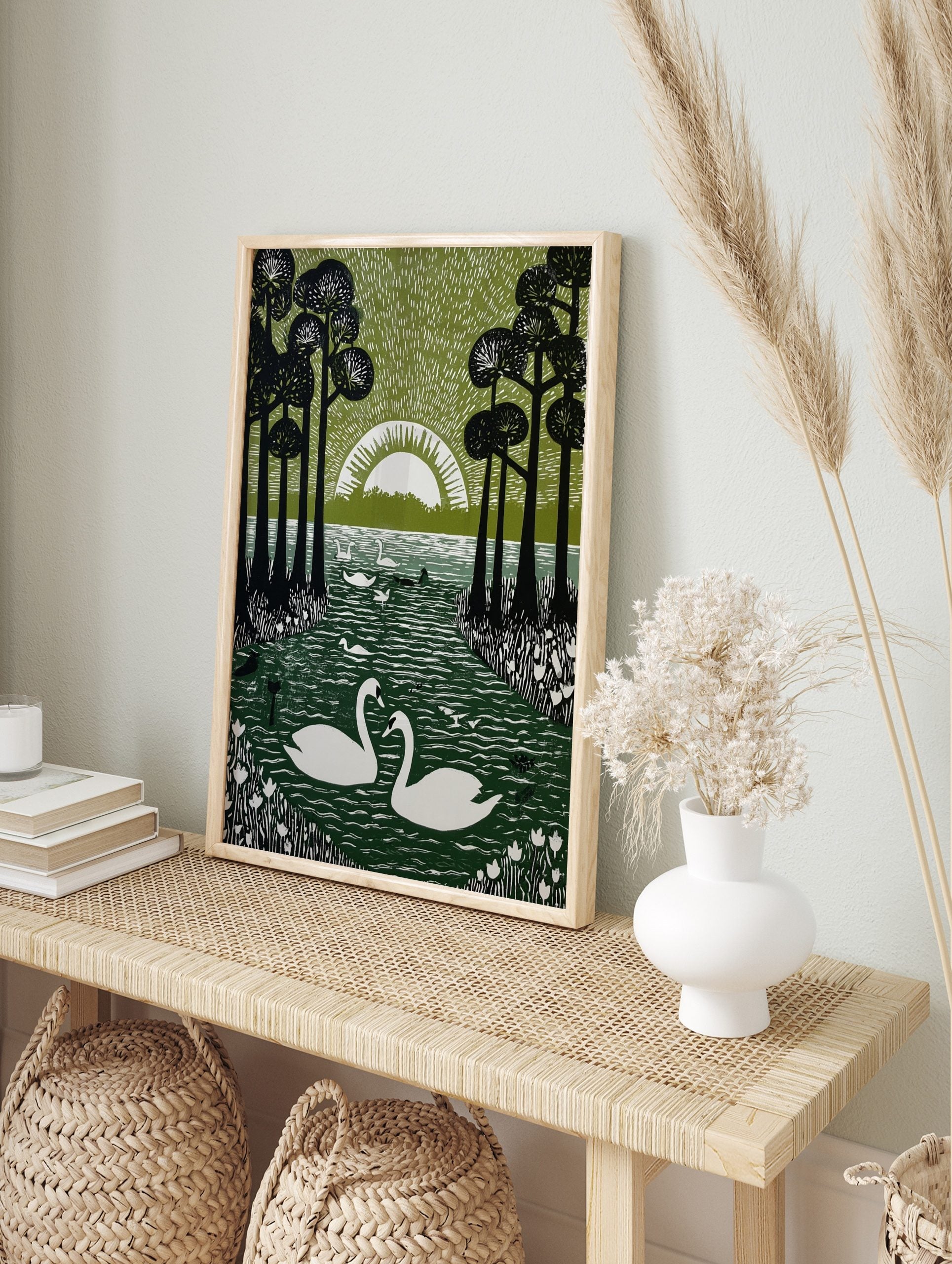 Linocut-style print of two white swans on a dark green lake, framed by tall black trees and a, in an alternate framed mockup