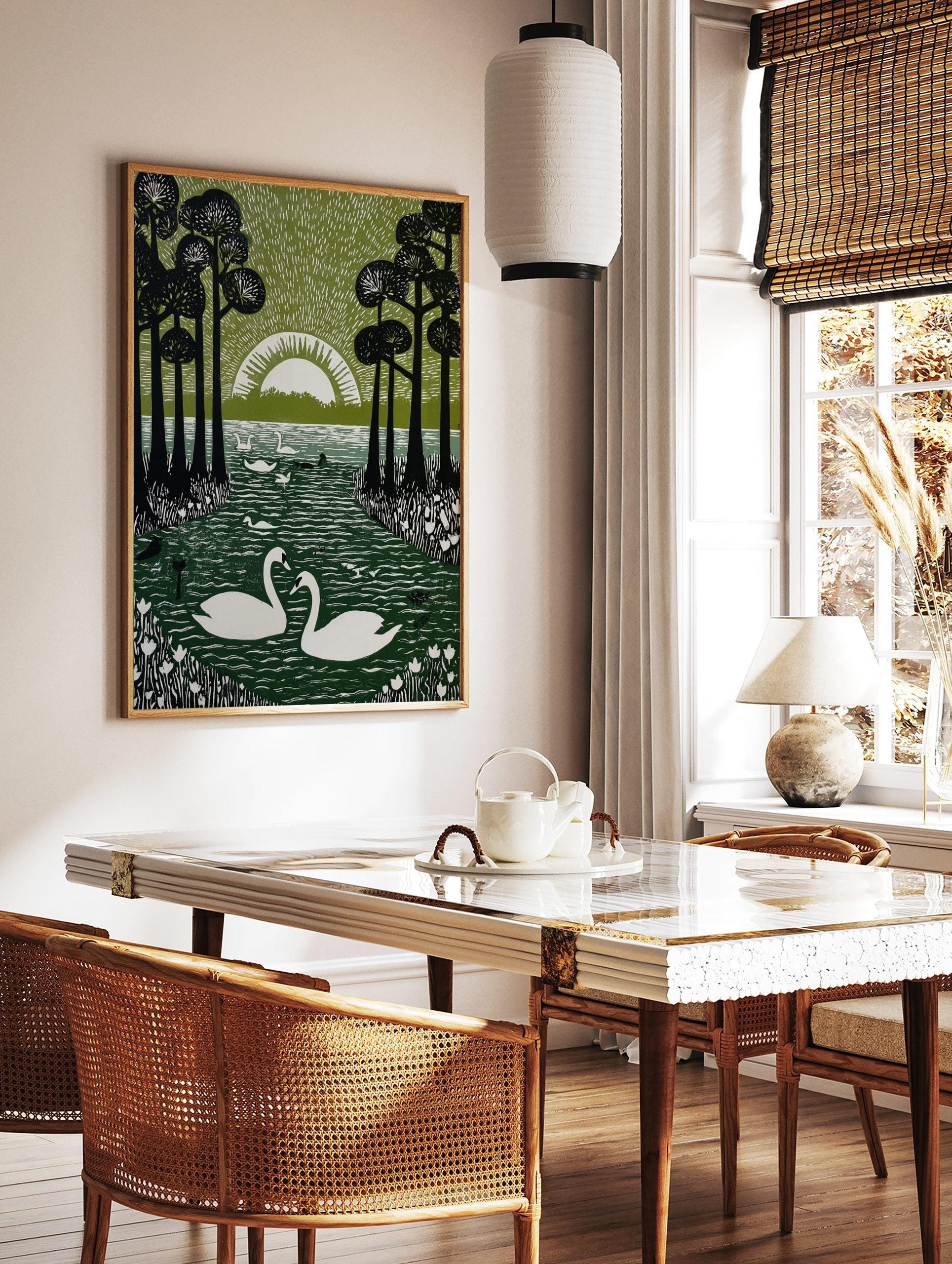 Linocut-style print of two white swans on a dark green lake, framed by tall black trees and a white, in a styled room mockup