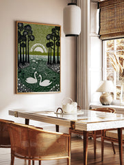 Linocut-style print of two white swans on a dark green lake, framed by tall black trees and a white, in a styled room mockup