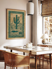 Teal saguaro cactus illustration in a desert landscape with hills and an ornate border, on tan paper, in a styled room mockup