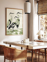 Minimalist illustration of three herons with black outlines and olive green bodies standing in water, in a styled room mockup