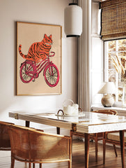 Orange tiger-striped cat riding a pink bicycle illustration on a textured beige background, in a styled room mockup