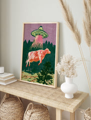 Retro illustration of a UFO abducting a cow with a pink tractor beam against a purple sky and, in an alternate framed mockup