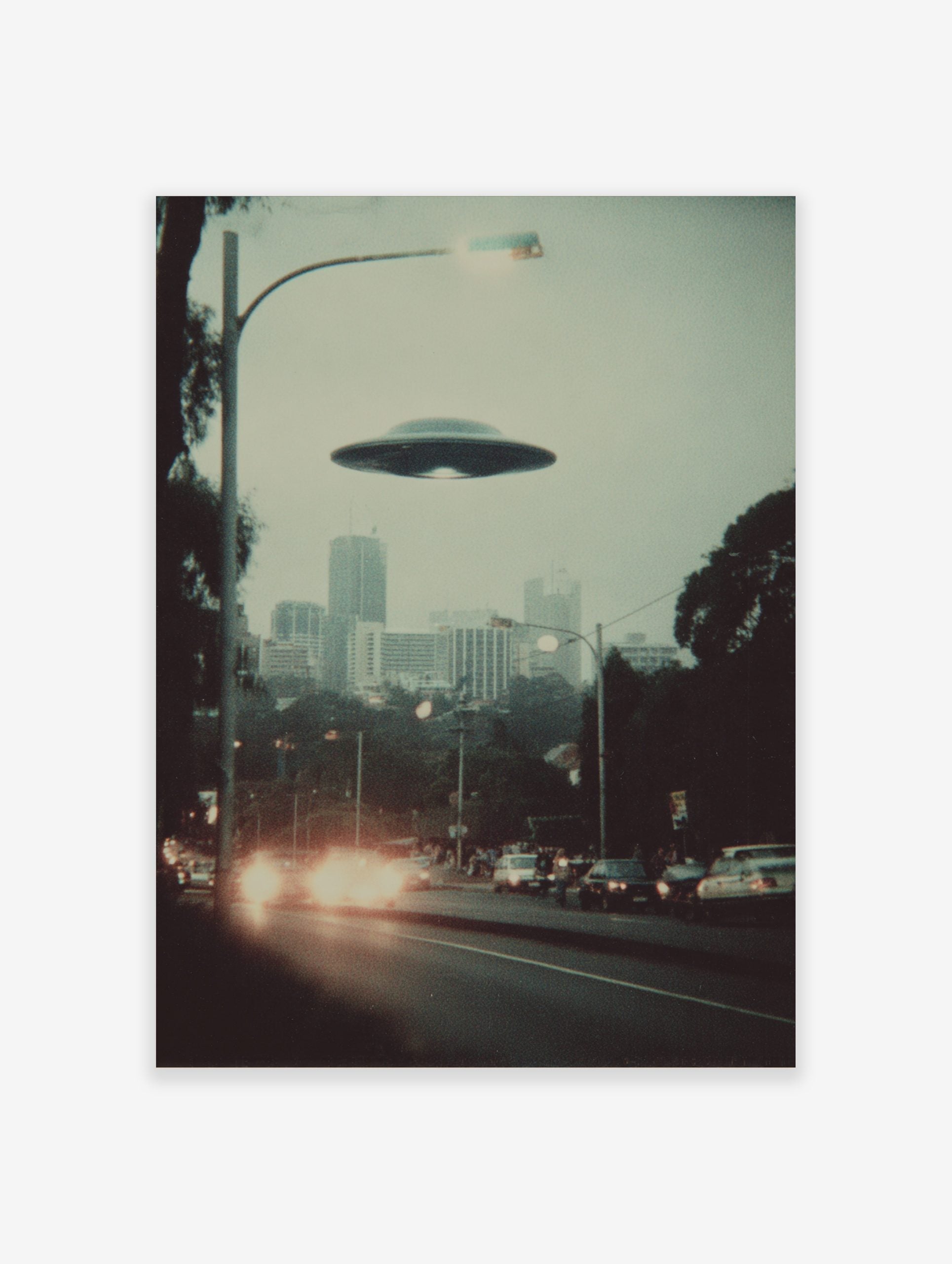 Retro photographic print of a flying saucer hovering over a hazy city street with car headlights and tall buildings