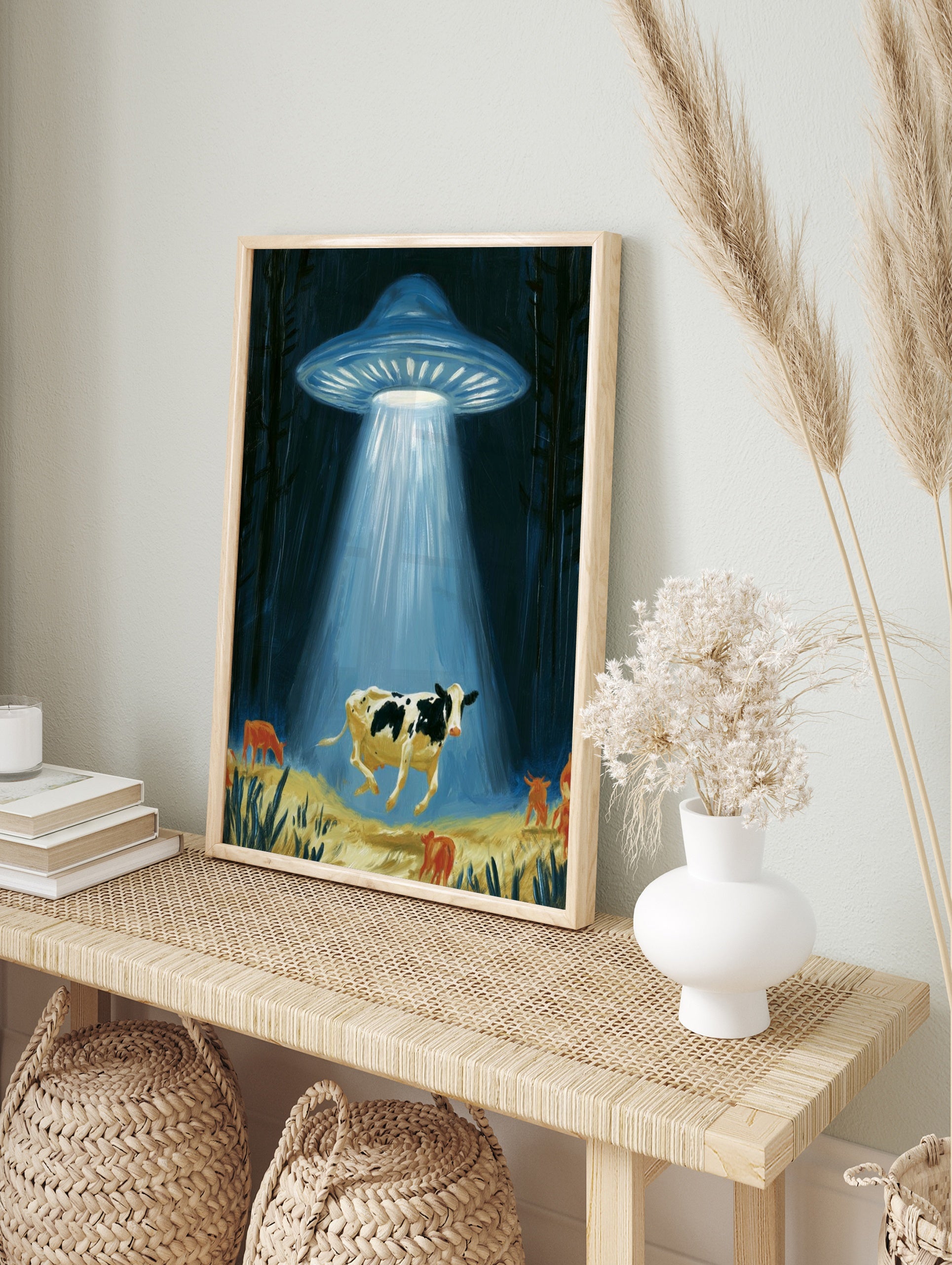 Surreal illustrative art print of a UFO abducting a cow with a glowing light beam in a dark, in an alternate framed mockup