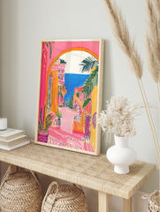 Vibrant illustrative print of a coastal town through an archway, featuring pink buildings, in an alternate framed mockup