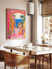 Vibrant illustrative print of a coastal town through an archway, featuring pink buildings, palm, in a styled room mockup