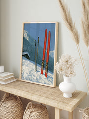 Vintage red skis and poles in deep snow with a mountain cabin and forest in the background, in an alternate framed mockup