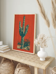 Illustrative Saguaro cactus print in dark teal and beige against a vibrant reddish-orange, in an alternate framed mockup