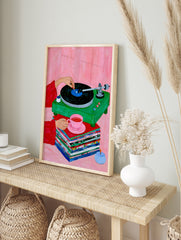 Illustration of a green vinyl record player, stacked records, and a pink teacup on a pink, in an alternate framed mockup