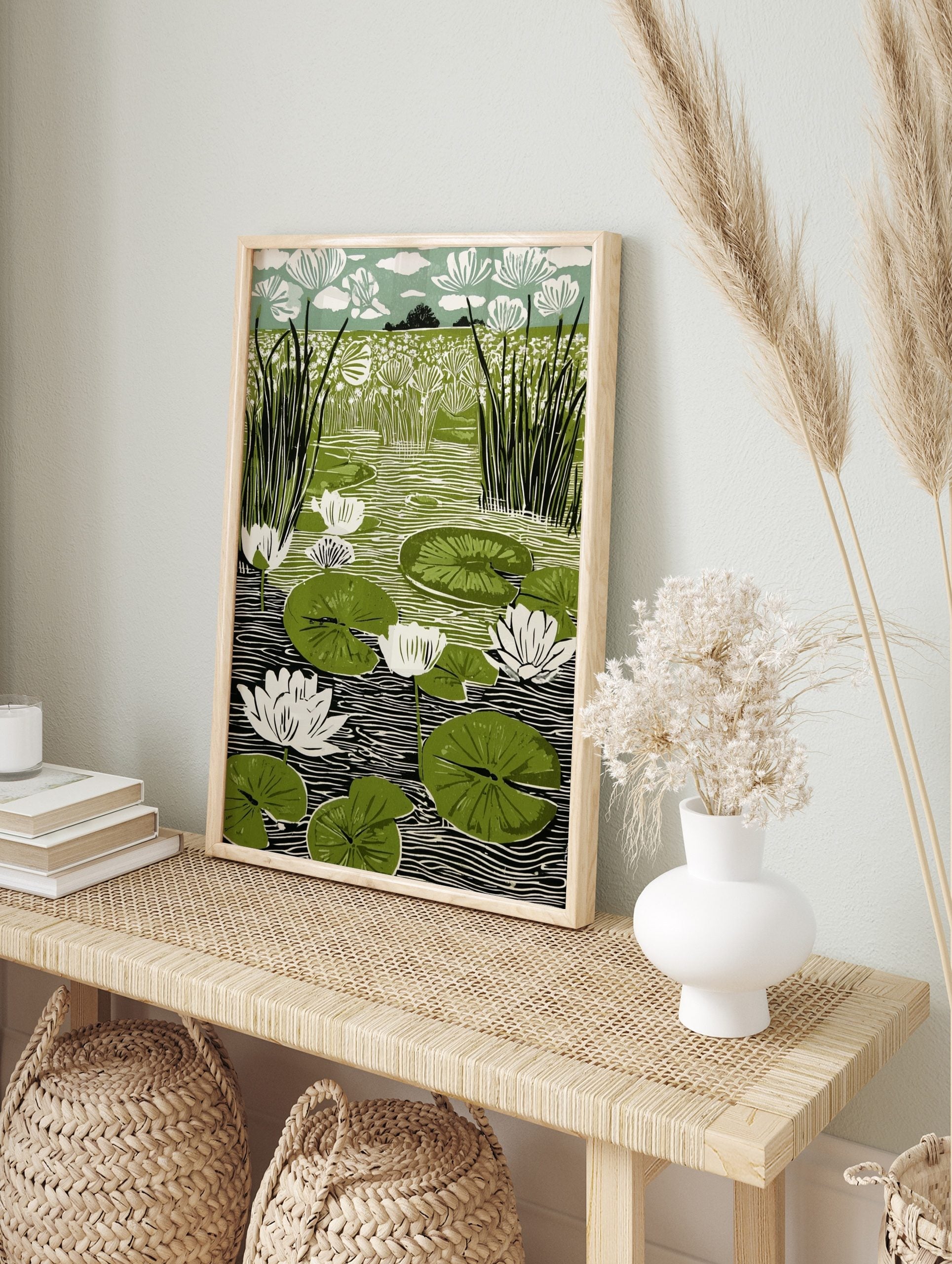 Linocut print of a pond with white water lilies, green lily pads, and tall reeds against a, in an alternate framed mockup