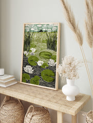 Linocut print of a pond with white water lilies, green lily pads, and tall reeds against a, in an alternate framed mockup
