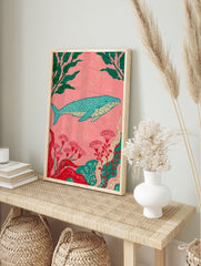 Stylized blue whale illustration with green kelp and red flowers on a pink background, in an alternate framed mockup