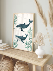 Two illustrative blue whales swimming horizontally amidst green coral branches on a white, in an alternate framed mockup