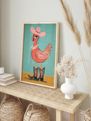 Whimsical chicken wearing pink cowboy hat, pearl necklace, and brown boots on teal and yellow, in an alternate framed mockup