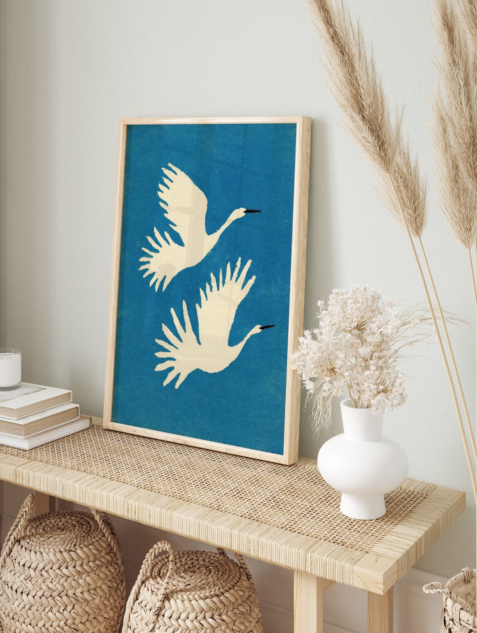 Two white cranes with black beaks flying upwards against a blue, textured background in a, in an alternate framed mockup