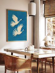 Two white cranes with black beaks flying upwards against a blue, textured background in a, in a styled room mockup