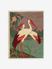 Two white cranes with red heads and wing tips standing in green water with foliage and pink flowers