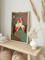 Two white cranes with red heads and wing tips standing in green water with foliage and pink, in an alternate framed mockup